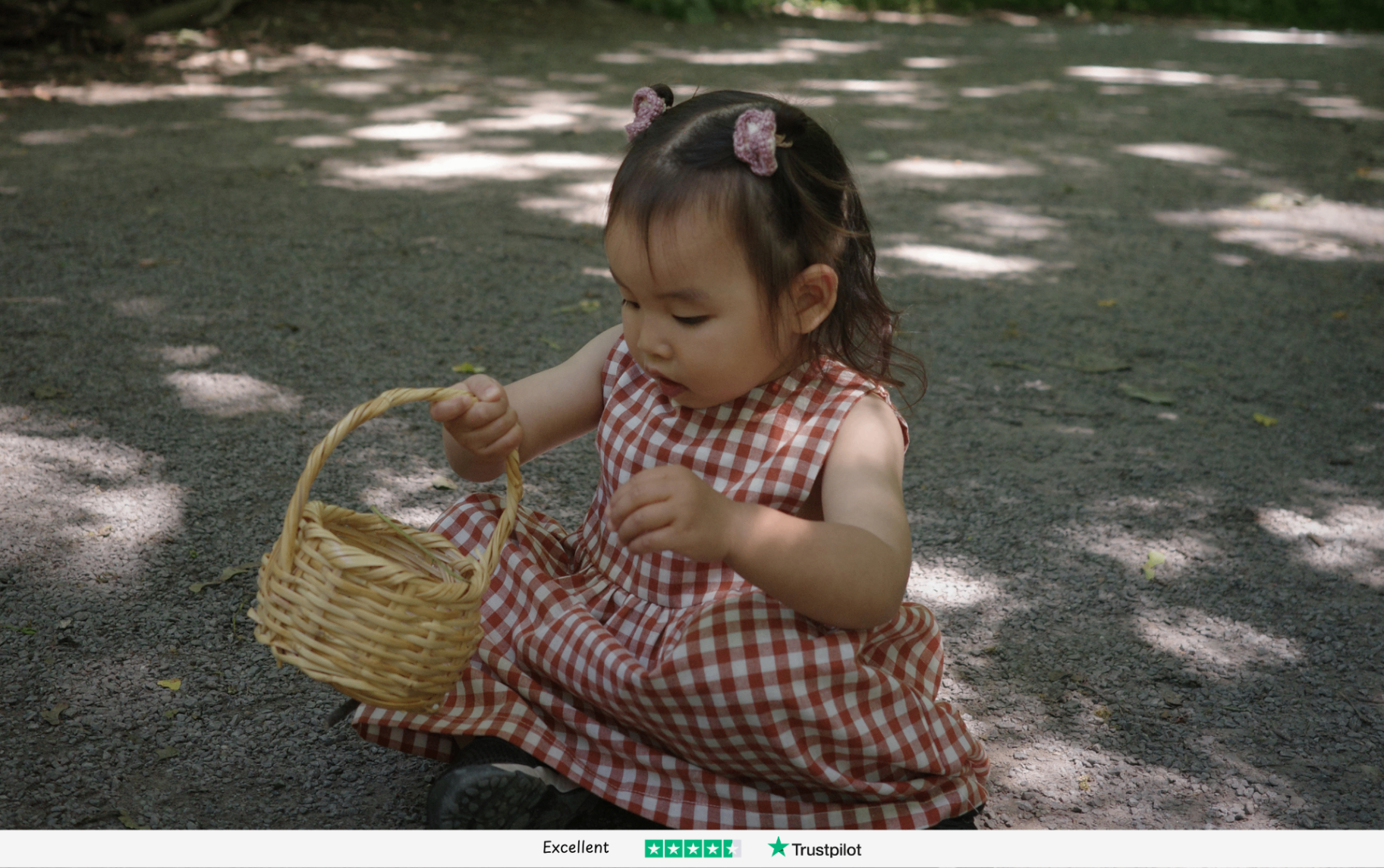 Toddler in a rust gingham dress holding a wicker basket on a sunlit woodland path - Little Novice children's clothing