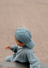 Child wearing a light blue cap and outfit sitting on a wooden surface with a beige background