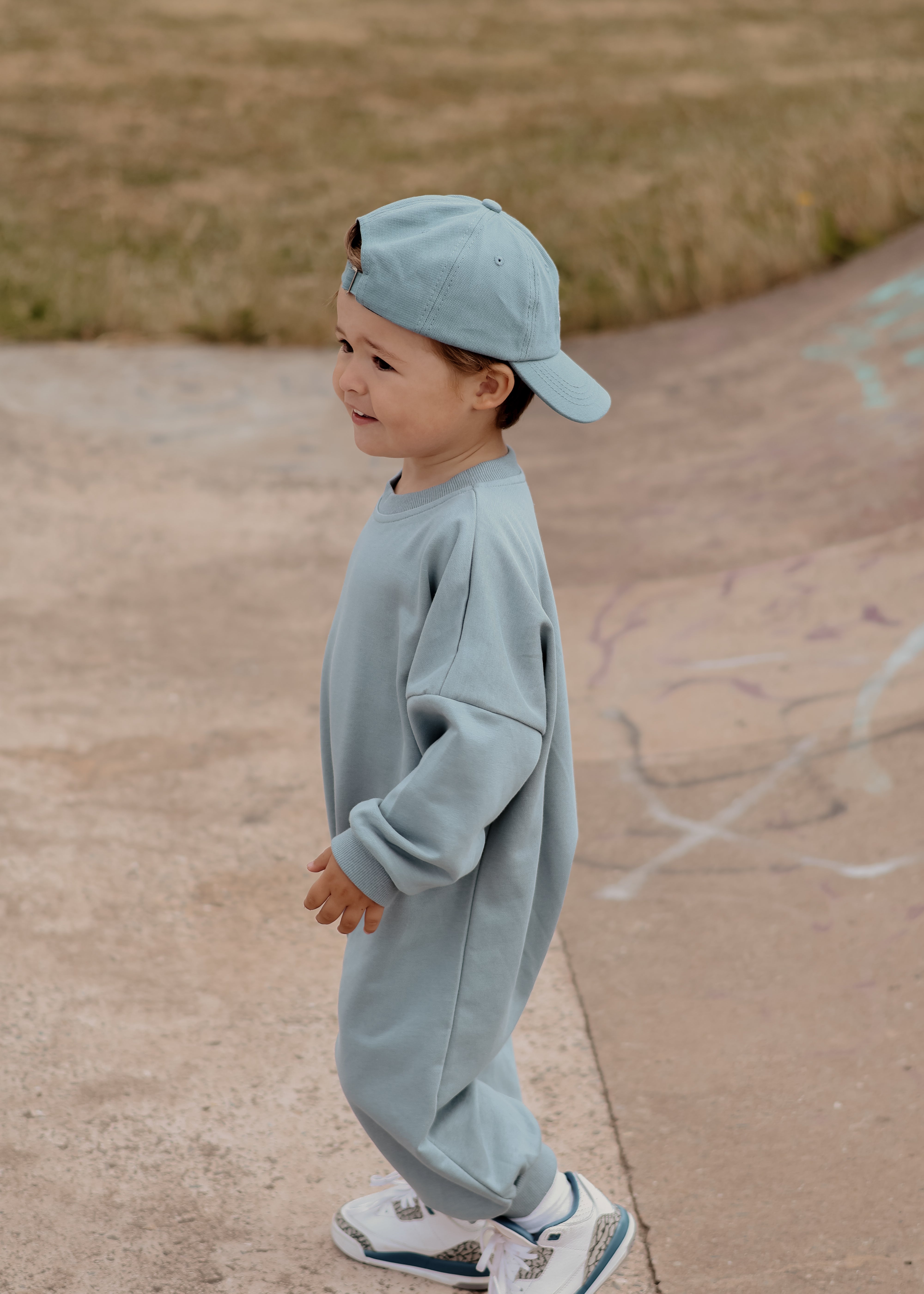 Child wearing a light blue outfit and cap standing on a concrete surface with grass in the background